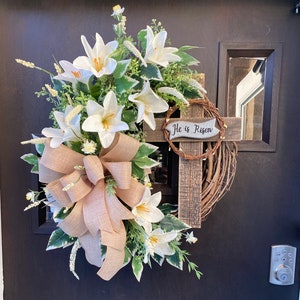 May include: A white lily wreath with a brown burlap bow and a wooden cross with the text "He is Risen" on a black door.