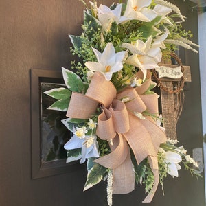 May include: A white and green Easter wreath with a burlap bow and a wooden sign that reads "He is Risen".