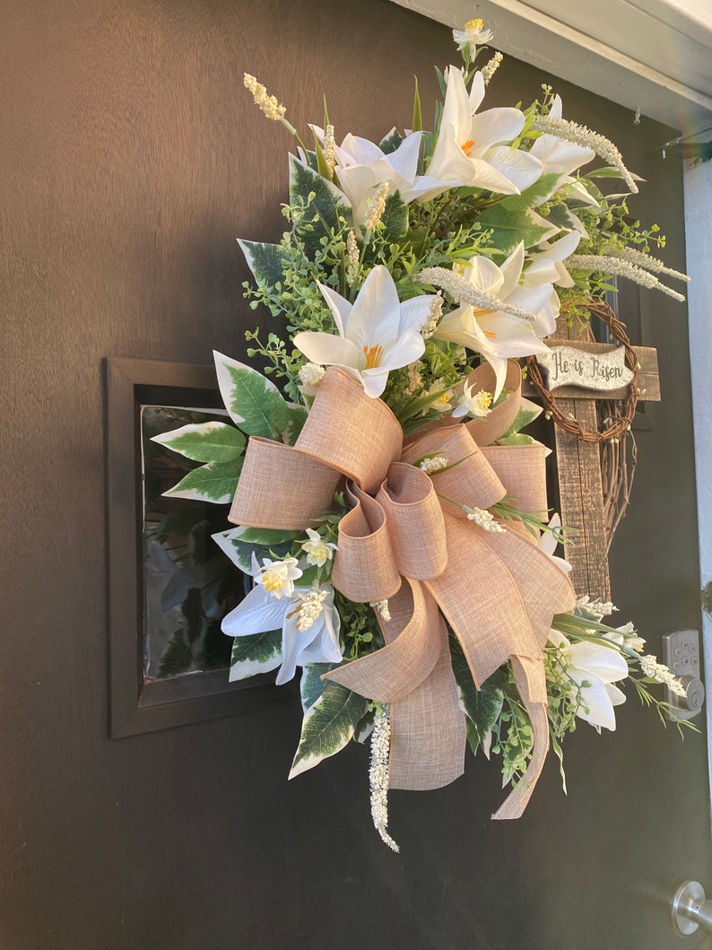 May include: A white and green Easter wreath with a burlap bow and a wooden sign that reads "He is Risen".