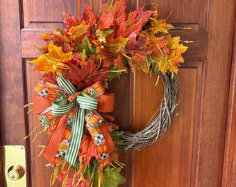May include: A fall-themed wreath with a grapevine base, adorned with colorful autumn leaves in shades of red, orange, and yellow. A large bow with pumpkin and striped ribbon accents the wreath, creating a festive seasonal decoration.