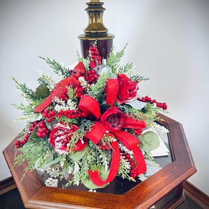 May include: A red and green Christmas floral arrangement with red roses, red berries, and a red ribbon sits on a wooden table with a glass top. A lamp with a white lampshade is in the background.