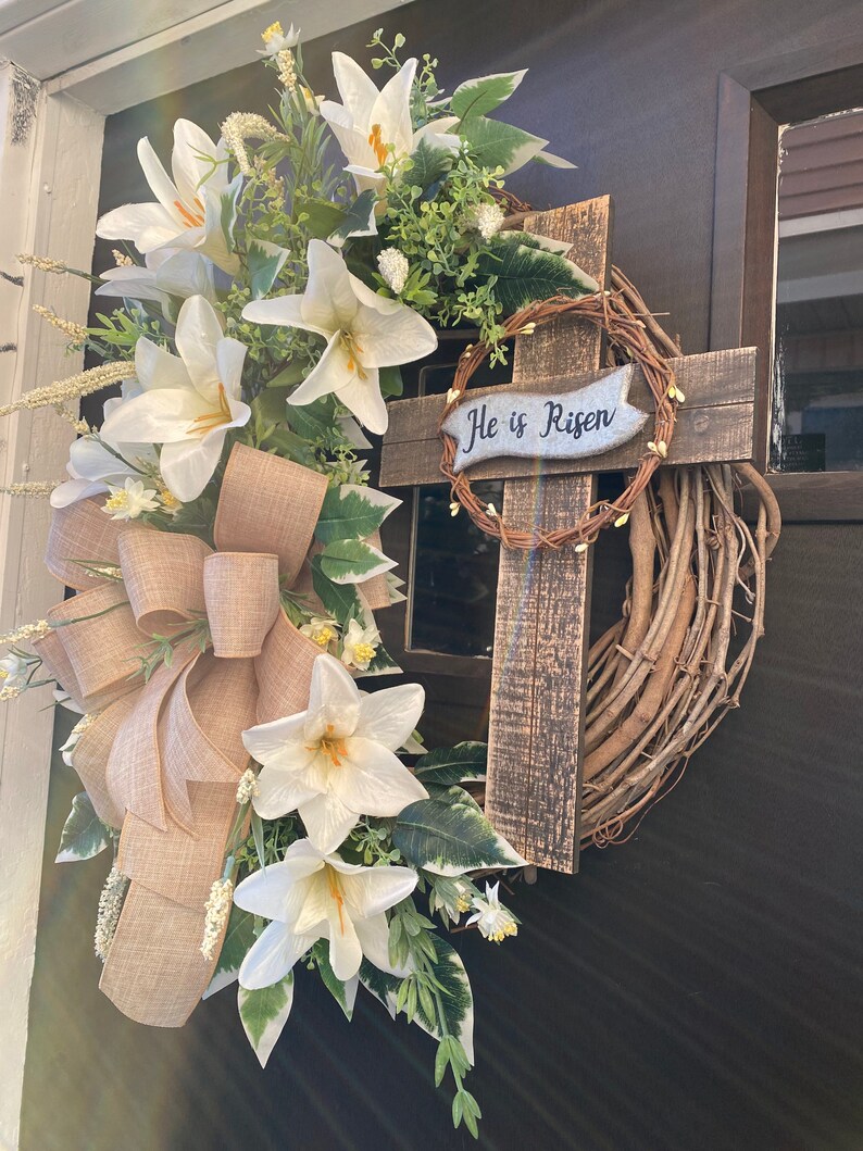May include: A white floral wreath with a wooden cross and the text "He is Risen" on a brown door.