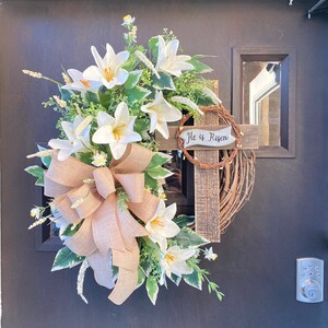 May include: A white and green Easter wreath with a burlap bow and a wooden cross with the text "He is Risen".