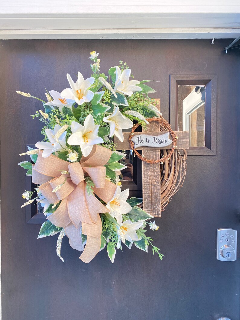 May include: A white and green Easter wreath with a burlap bow and a wooden cross with the text "He is Risen".