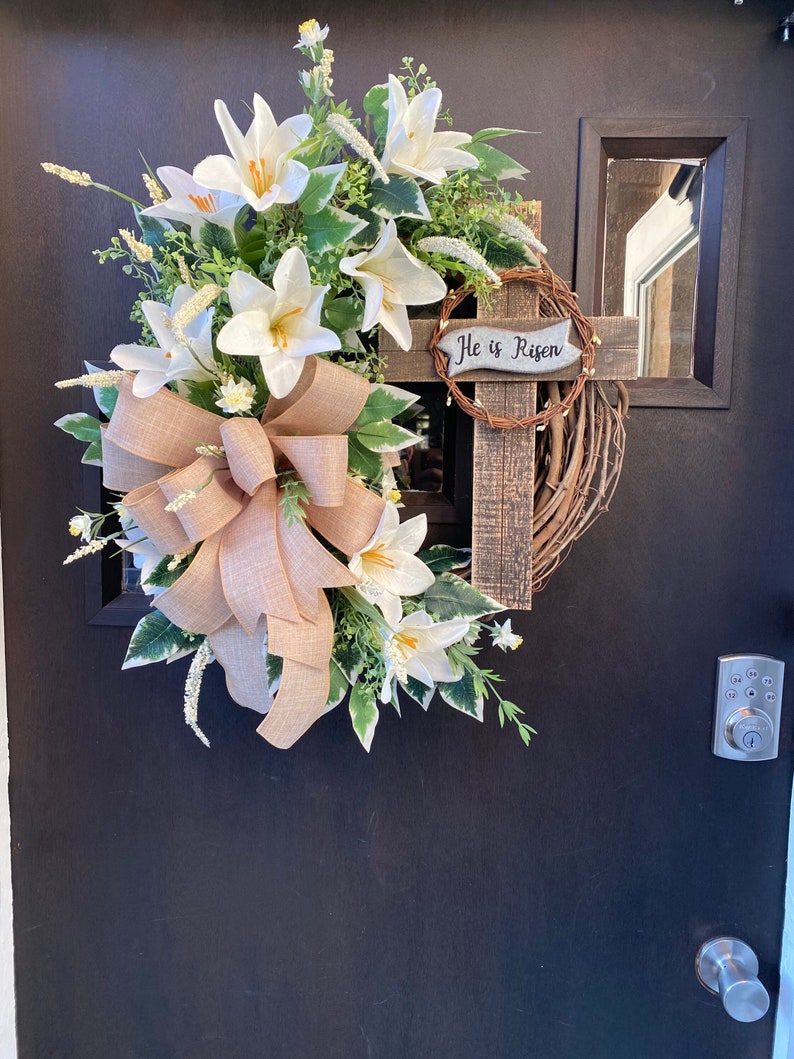 May include: A white lily wreath with a brown burlap bow and a wooden cross with the text "He is Risen" on a black door.