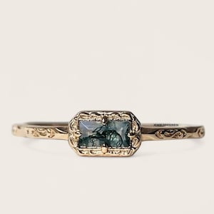 Puede incluir: Un anillo de oro con una piedra de ágata verde y blanca engastada en un marco ornamentado y detallado. La banda del anillo es fina y tiene un delicado diseño floral.
