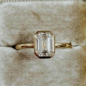 May include: A gold ring featuring an emerald-cut diamond. The diamond is set in a bezel setting, and the band is slender. The ring is presented in a grey velvet ring box.