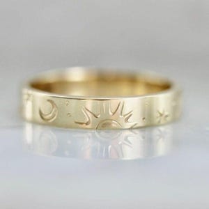Puede incluir: Un anillo dorado con grabados celestiales. El anillo presenta un diseño de sol, luna y estrella alrededor de la banda. El anillo mide aproximadamente 0,6 cm de ancho y tiene un acabado pulido, que refleja la luz.