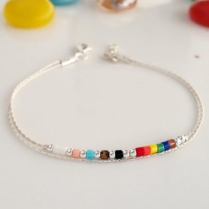 Può includere: Un braccialetto a catena d'argento con una fila di perline colorate. Le perline sono disposte a forma di arcobaleno, con perline bianche, pesca, turchese, marroni, nere, argentate, rosse, arancioni, gialle, verdi, blu e viola.