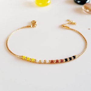 Könnte beinhalten: Ein filigranes goldfarbenes Armband mit einer Reihe kleiner, farbiger Perlen. Die Perlen sind gelb, weiß, rotbraun und schwarz. Das Armband hat ein kettenartiges Design und einen Verschluss. Ein stilvolles Schmuckstück.