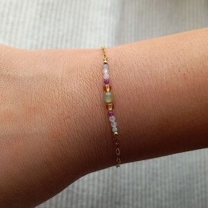 May include: A delicate gold chain bracelet featuring a row of colourful gemstone beads. The beads are in shades of pink, blue, green, and yellow.
