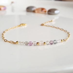 May include: A delicate gold chain bracelet featuring a row of small, faceted gemstone beads in shades of white, pink, and blue.