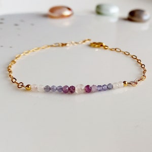 May include: A delicate gold chain bracelet featuring a row of small, colourful gemstone beads. The beads are arranged in a gradient pattern, starting with white, then light blue, purple, and ending with white again.