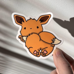 Puede incluir: Una pegatina con un personaje de dibujos animados Eevee en tonos naranja, marrón y blanco. Eevee tiene una expresión linda y detalles en forma de corazón en sus pies. La pegatina está sobre un fondo blanco.