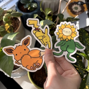 Puede incluir: Tres pegatinas coloridas con personajes de dibujos animados. Uno es una criatura naranja con forma de zorro, otro es un personaje amarillo con mejillas rojas y el tercero es un girasol sonriente con hojas y tallo verdes.