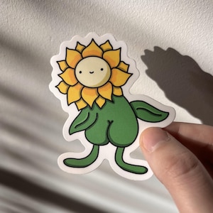 Puede incluir: Una pegatina caprichosa con un personaje de girasol de dibujos animados. El girasol tiene pétalos amarillos, un centro color crema y una cara sonriente. El tallo y las piernas son verdes, y la pegatina está sujeta por una mano.