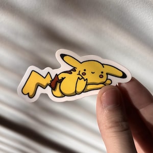 Puede incluir: Una pegatina de Pikachu amarilla con un contorno negro. La pegatina representa a Pikachu acostado con los ojos cerrados y una expresión relajada. La cola es amarilla con una base marrón. La pegatina está sujeta por una mano.