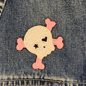 May include: A pink and white embroidered skull patch with black accents. The patch is sewn onto a blue denim jacket.
