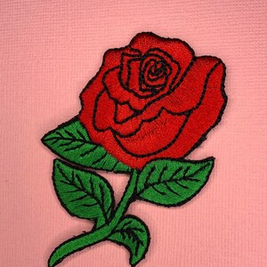 May include: An embroidered red rose with green leaves and a stem. The rose is on a pink background.