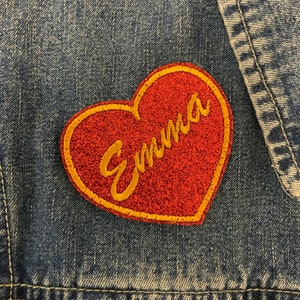 May include: Red heart-shaped patch with a yellow outline and the word "Emma" written in yellow cursive letters. The patch is sewn onto a blue denim jacket.