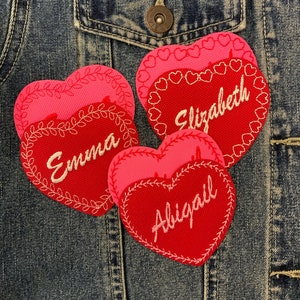 May include: Three red felt heart-shaped patches with pink outlines and white embroidery. The patches are sewn onto a blue denim jacket. The patches are personalised with the names "Emma", "Abigail", and "Elizabeth".