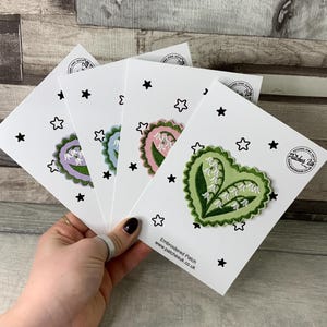 May include: Four white cards, each with a heart-shaped embroidered patch featuring a lily of the valley design. The patches are in shades of purple, blue, pink, and green. Black stars and star shapes surround the patches. The text "Embroidered Patch" and a website address are visible.