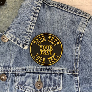 May include: A black and yellow round patch with the text "YOUR TEXT" repeated three times. The patch is sewn onto a blue denim jacket.