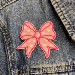 May include: A pink bow with white hearts embroidered on it. The bow is iron-on and can be used to decorate clothing, bags, or other items.