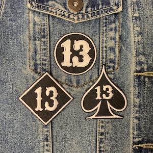 May include: Three black fabric patches with white stitching and the number "13" on a denim jacket. The patches are a circle, a diamond, and a spade.