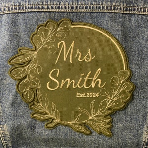 May include: A green embroidered patch with a floral wreath design. The patch says "Mrs. Smith Est. 2024" in a cream coloured font.