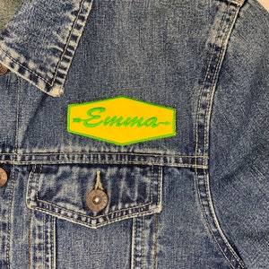 May include: A denim jacket with a yellow and green embroidered patch that says "Emma" on it.