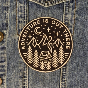 May include: A black and white embroidered patch with the text "Adventure is out there" and a design of a campfire, mountains, and trees. The patch is on a blue denim jacket.