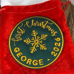 May include: A red velvet drawstring pouch featuring a circular dark green embroidered patch. The patch displays a gold snowflake in the centre, surrounded by the yellow embroidered text "First Christmas" and "GEORGE - 2026".