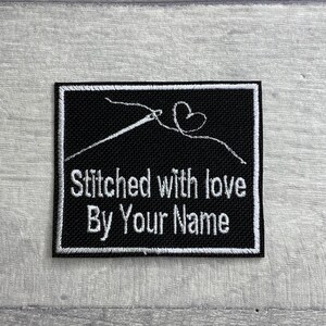 May include: Black and white embroidered patch with a needle and thread forming a heart. The text reads "Stitched with love By Your Name".