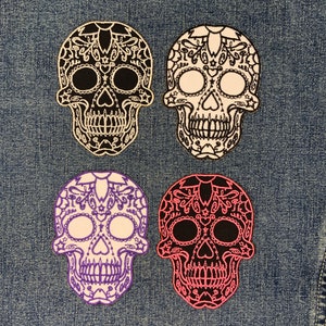 May include: Four sugar skull patches in different colours. The skulls are black, white, pink, and purple. Each skull has intricate designs and details.