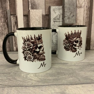 May include: Two white ceramic mugs with black rims. The mug on the left has a black handle and features a skull with a crown and beard, roses, and the word "Mr". The mug on the right has a black handle and features a skull with a crown and roses, and the word "Mrs".