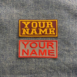 May include: Two embroidered patches with the text "YOUR NAME" in yellow and red lettering. The top patch has a maroon background with a yellow border. The bottom patch has a grey background with a red border.