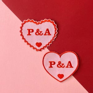 May include: Two heart-shaped patches with red embroidered trim and lettering. The patches are white with the letters "P & A" in red, and a small red heart at the bottom. One patch has a scalloped edge, set against a red and pink background.