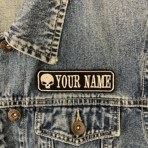 May include: A black and white embroidered patch with a skull and the text "YOUR NAME" on a denim jacket.