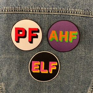May include: Three round fabric patches with different letters and colours. The first patch is white with red letters that spell "PF". The second patch is purple with green letters that spell "AHF". The third patch is black with pink and yellow letters that spell "ELF".