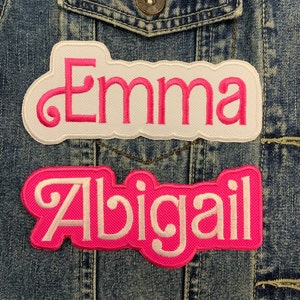 May include: Two white fabric patches with pink lettering sewn onto a blue denim jacket. The top patch says "Emma" and the bottom patch says "Abigail".