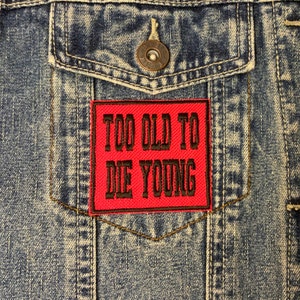 May include: A red denim patch with black embroidered text that reads "Too Old To Die Young" on a blue denim jacket.