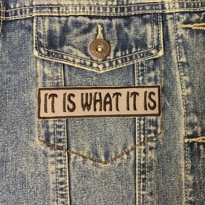 May include: A denim jacket with a brown patch sewn onto the pocket. The patch has white text that reads "IT IS WHAT IT IS".