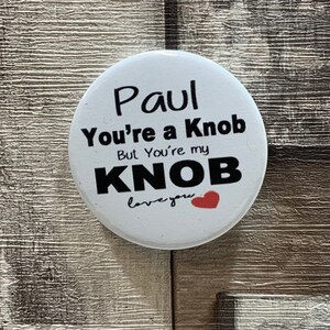 May include: White button with black text that reads "Paul You're a Knob But You're my KNOB love you" with a red heart.