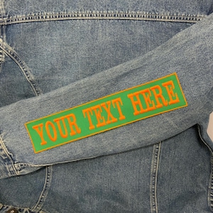 May include: A light blue denim jacket with a green rectangular patch on the sleeve. The patch has the text "YOUR TEXT HERE" in orange. The jacket is a classic style.