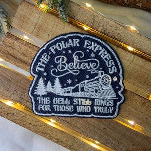 May include: A blue embroidered patch with the text "The Polar Express Believe The Bell Still Rings For Those Who Truly" and a white silhouette of a train with snow falling.