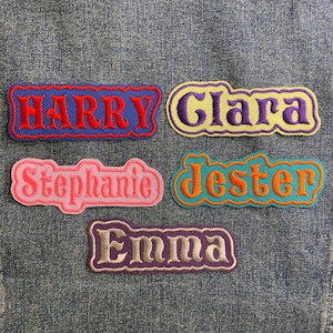 May include: Five embroidered name patches: "Harry" in red on blue, "Clara" in purple on yellow, "Stephanie" in pink, "Jester" in orange and teal, and "Emma" in white on purple. The patches are on a denim background.