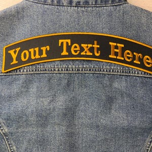 May include: A denim jacket with a black patch embroidered with the words "Your Text Here" in gold lettering. The patch is arched and sewn onto the back of the jacket.