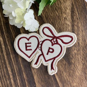 May include: An embroidered patch featuring two cream-coloured hearts outlined in dark red thread. The left heart contains the letter E and the right heart contains the letter P. A dark red ribbon design connects the two hearts at the top.