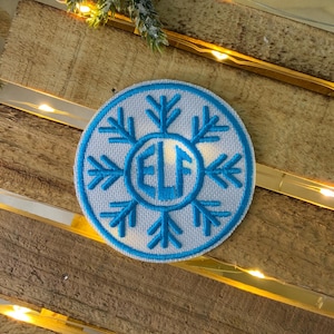 May include: A white, round, embroidered patch with a blue snowflake design. The snowflake has a circle in the centre with the letters "ELF" in blue.